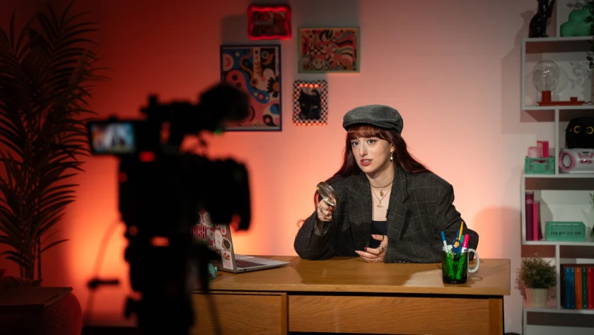 Eine junge Frau sitzt als Detektivin verkleidet in einem Fernsehstudio, das eingerichtet ist wie ein Wohnzimmer, an einem Schreibtisch und spricht in eine Kamera.